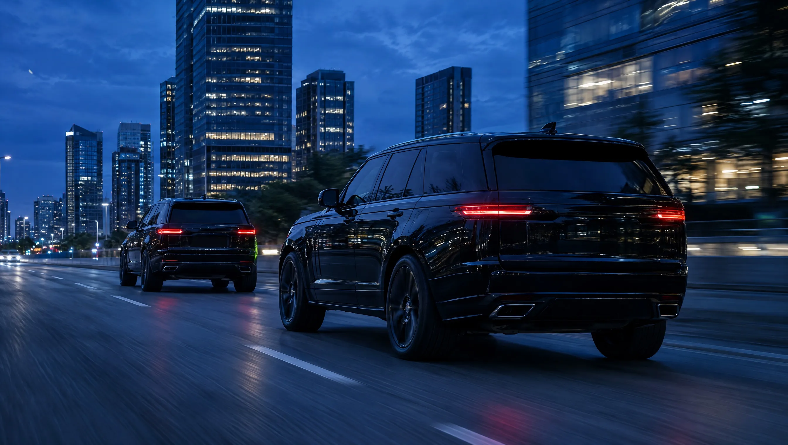 Two black SUVs travelling in convoy along a city avenue at blue hour
